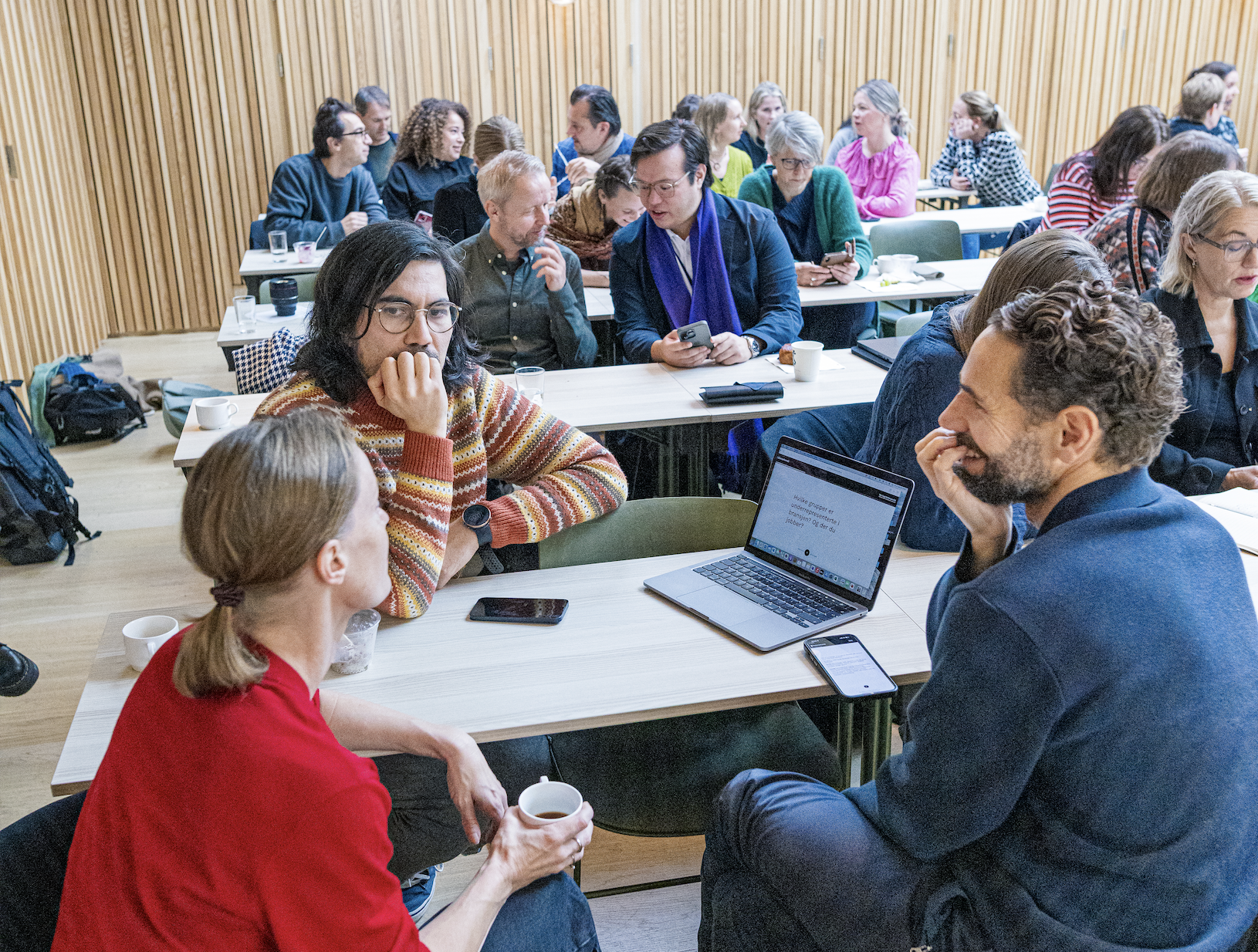 Photo of many people sitting in groups talking. Laptops on the table.