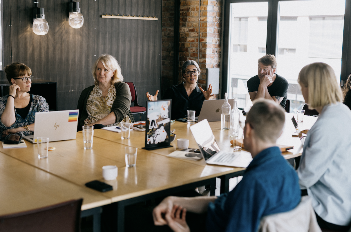 Foto av folk som snakker sammen rundt et bord. En person gestikulerer og de andre lytter.